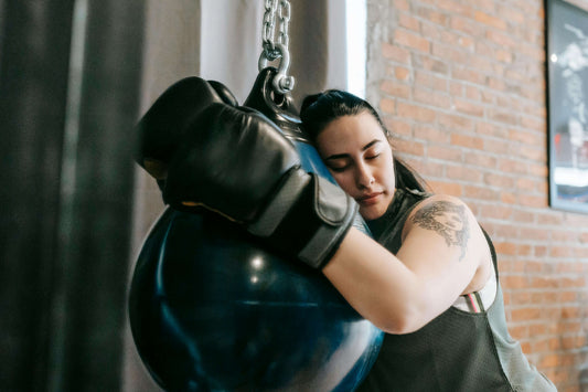 A woman in boxing gloves resting her head against a heavy bag, symbolising emotional release and the mental health benefits of boxing training.