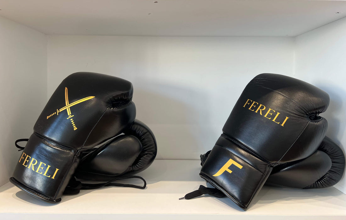 Black Fereli lace-up boxing gloves with gold logo and emblem displayed side by side on a white shelf.